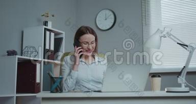 年轻的休闲女士坐在办公桌前，用笔记本电脑，接电话，打电话，微笑着在手机上说话